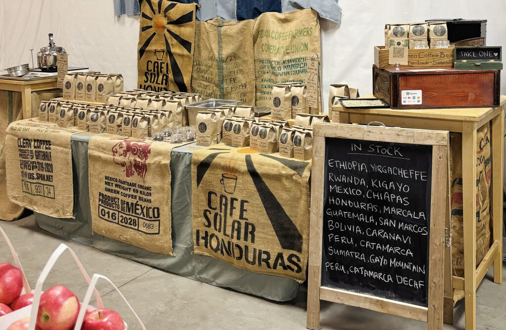 A full view of the She Grows Coffee booth at the St. Lawrence Market. Neatly packed and arranged coffee in kraft bags, on a table decorated in burlap coffee bags, and with a chalkboard listing the current beans in stock.